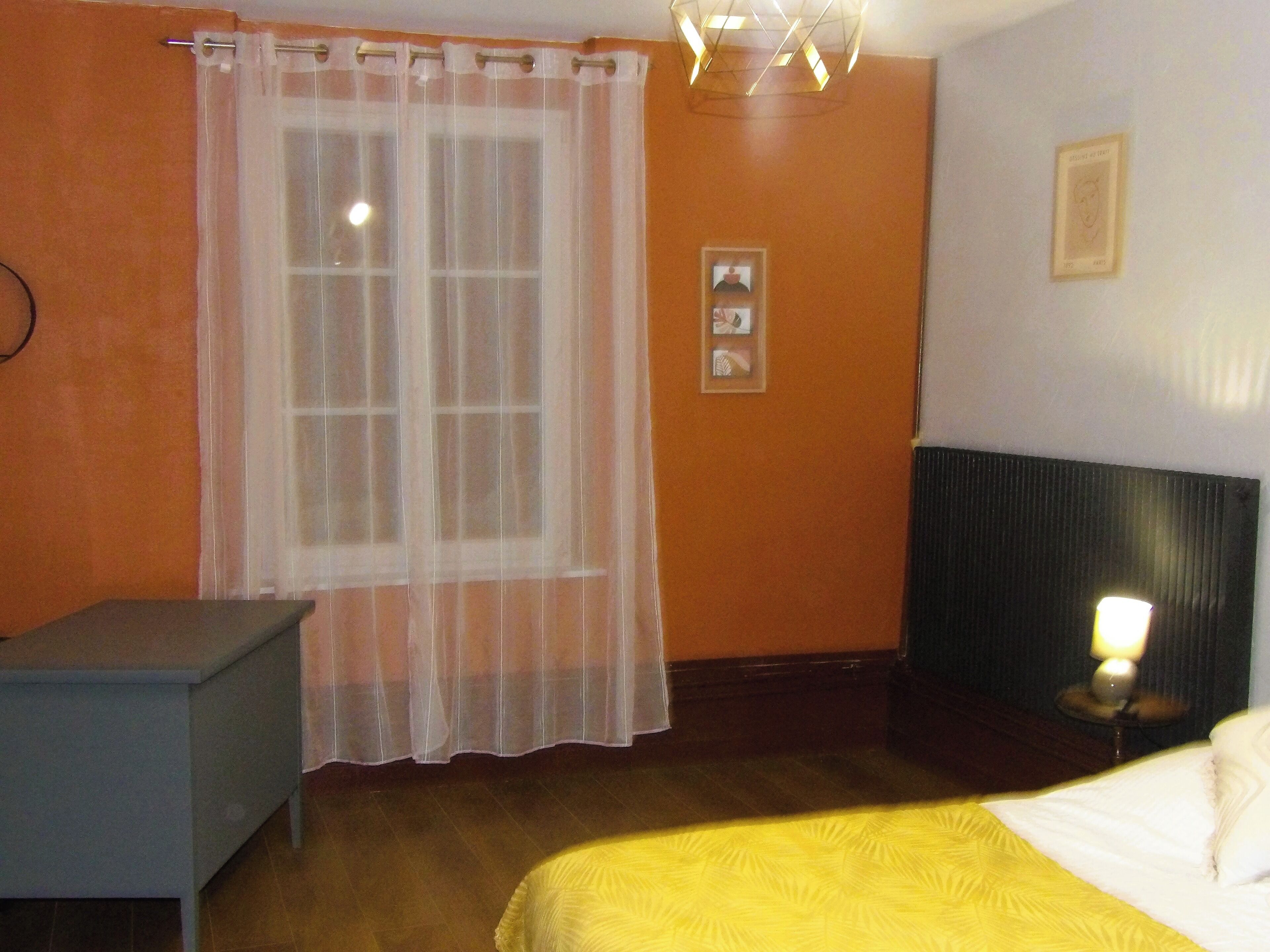 Habitación de hotel con cama, mesita de noche, ventana con cortinas translúcidas, un cuadro en la pared y una mesita con lámpara.