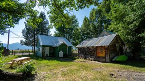 En rustik stuga med ett grönt tak och en träkonstruktion, omgiven av träd och ett picknickbord.