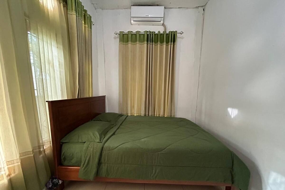 A single bed with a wooden frame and green bedding, a white air conditioning unit above, and a window with curtains.