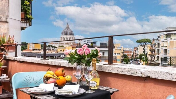 Une table dressée avec des fruits, un vase de fleurs et des verres à vin sur un balcon donnant sur une vue panoramique de la ville, avec ses bâtiments d’architecture historique.