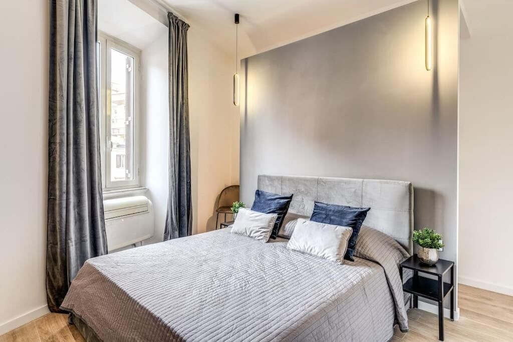 A modern bedroom with a large bed, grey curtains, a bedside table with a plant, and a wall-mounted light fixture.