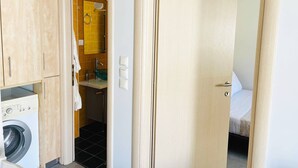 A compact room with a washing machine, a sink with a glass basin, and a bed with a white cover. - Cozy Nest - 2min To Beach, 6min Walk To (Chania)