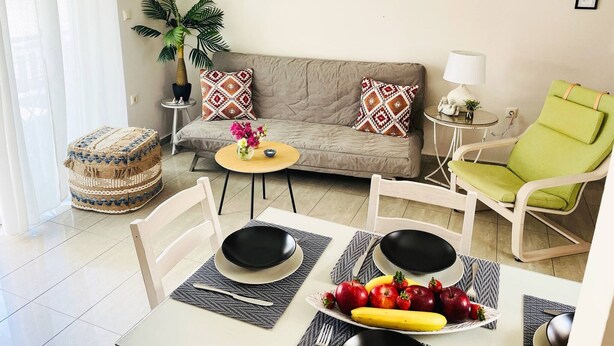 A living room with a sofa, a green chair, a round table with a bowl of fruit, and a small side table with a lamp. - Cozy Nest - 2min To Beach, 6min Walk To (Chania)