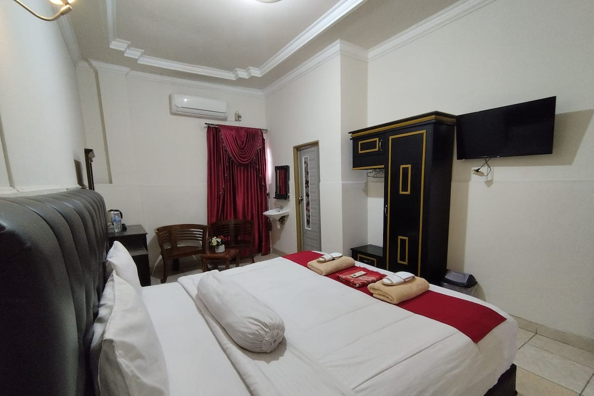 A hotel room with a bed, a desk, a chair, a wardrobe, a TV, and an air conditioning unit.