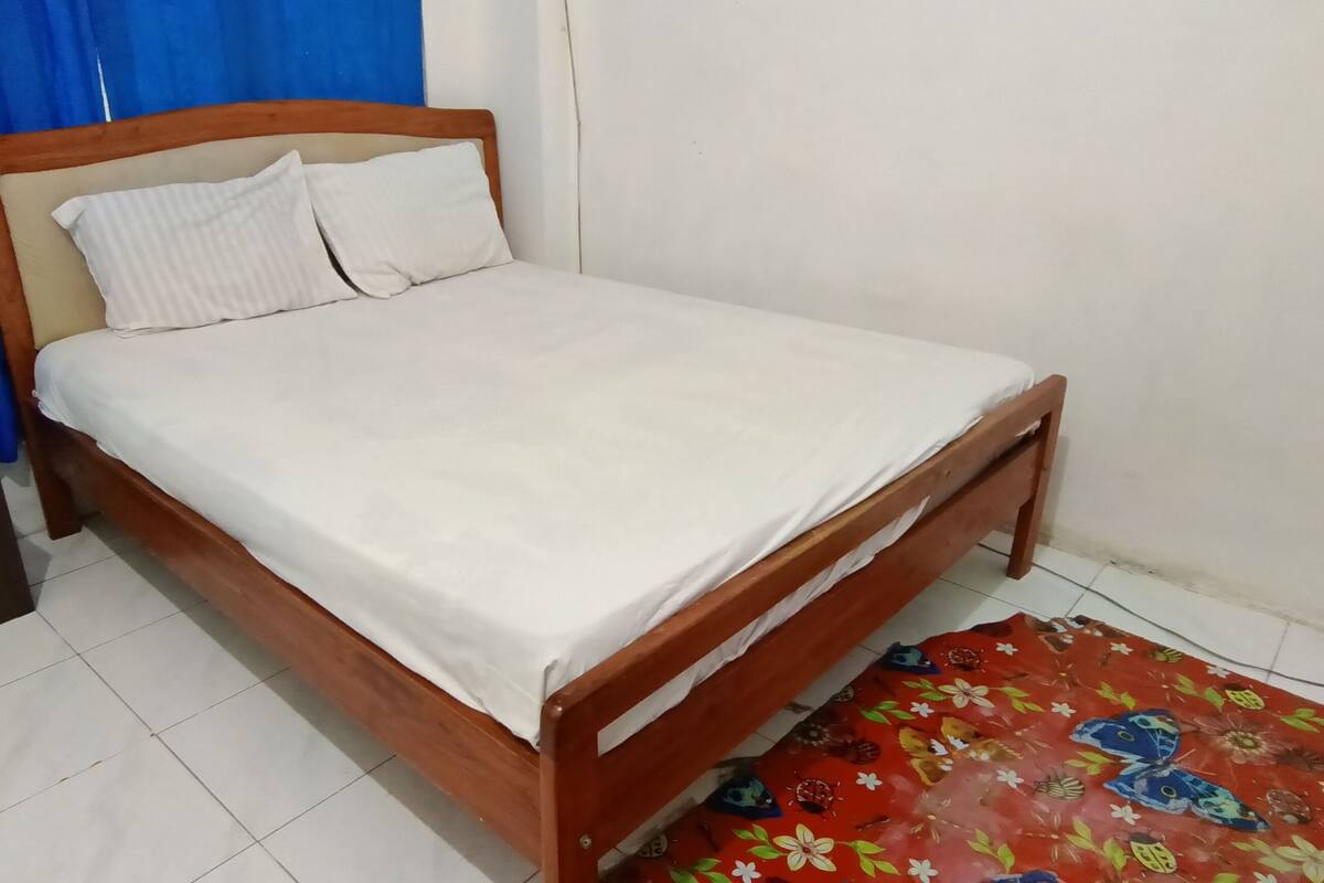 A single bed with a wooden frame and white bedding in a room with a tiled floor and a red floral rug.