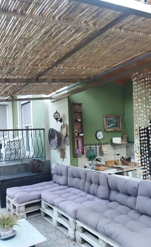 A cozy outdoor seating area with a cushioned sofa and a dining table, surrounded by various decorative items and a wooden ceiling.