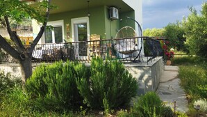 Edifici de dues plantes amb façana verda, un balcó amb barana metàl·lica i una zona de jardí amb arbustos i una planta en test.