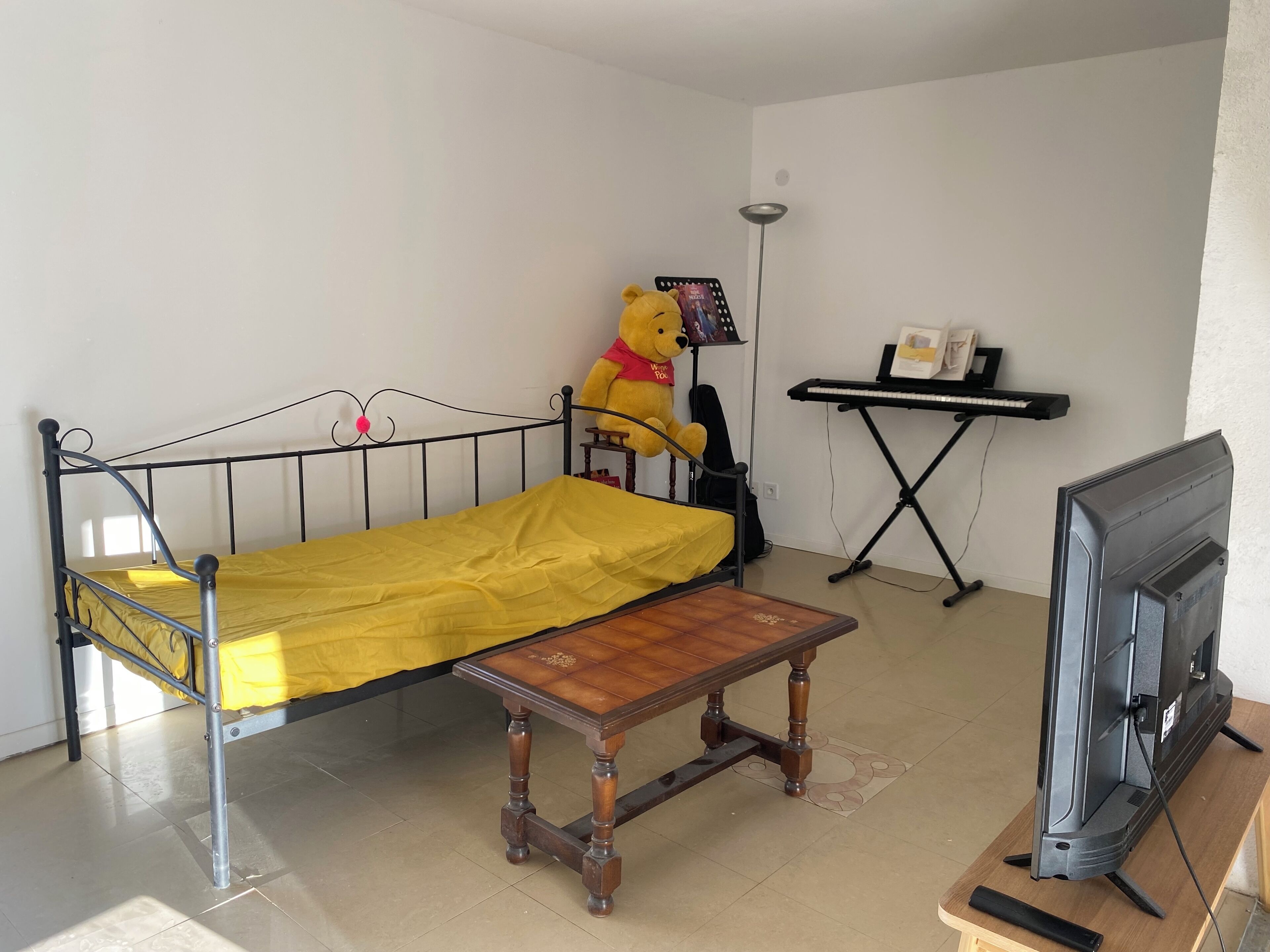 Ein kleines Zimmer mit einem Metallbettgestell, einer gelben Matratze, einem Holztisch, einem Fernseher und einem Teddybär.