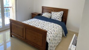 A bedroom with a wooden bed, a nightstand, a window with a view of a parking lot, and a white air conditioning unit. - Petite Maison Avec Terrain Entre Route, Vignes et Lavande (Sainte-Cécile-les-Vignes)