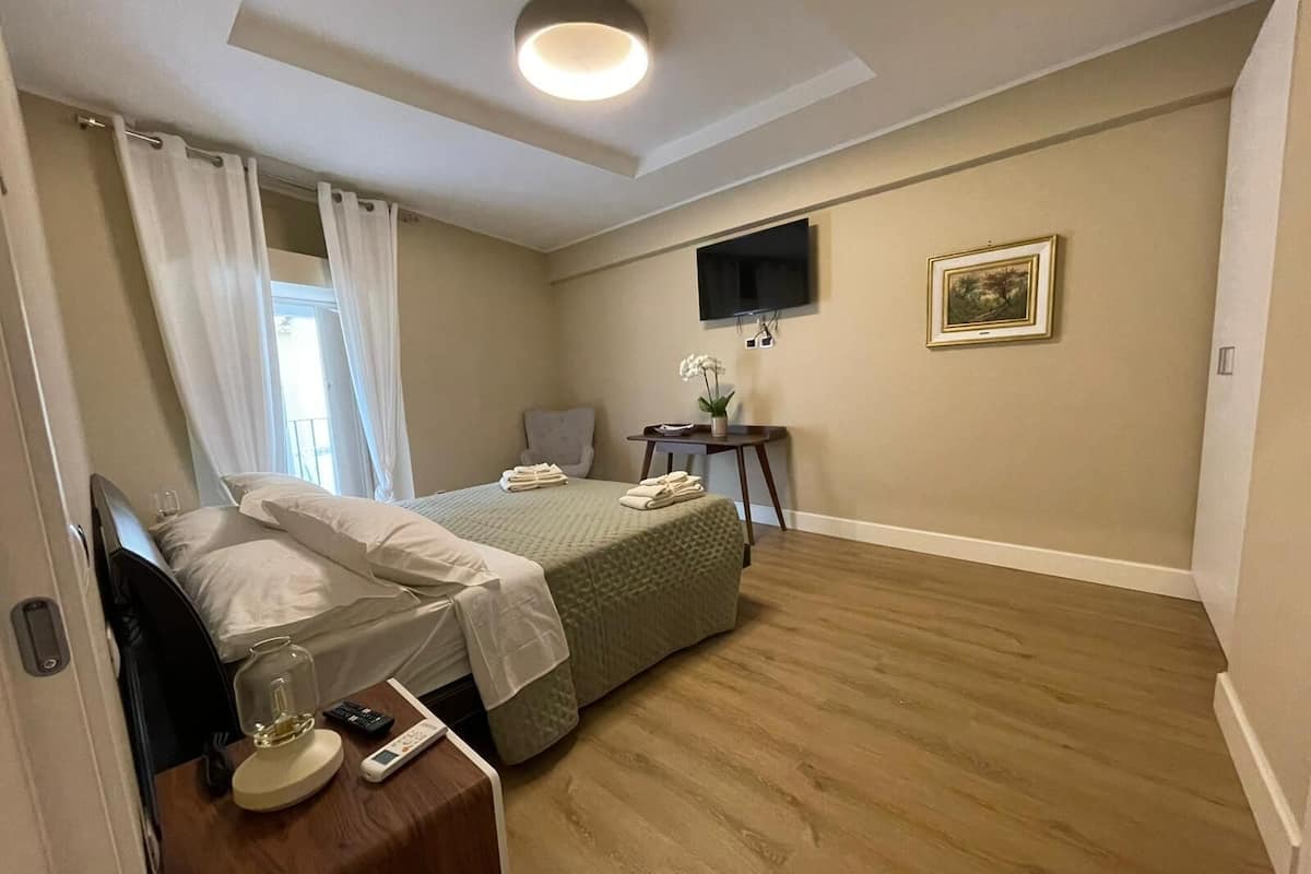 A hotel room with a bed, bedside table with a lamp, a small table with a vase of flowers, a chair, a TV on the wall, and a framed picture.