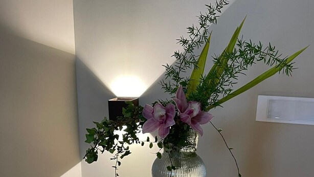 A tall, narrow vase with a textured surface containing pink flowers and green foliage, placed against a plain wall with a light fixture above it. - Ai Cortili  (Sant'Egidio del Monte Albino)