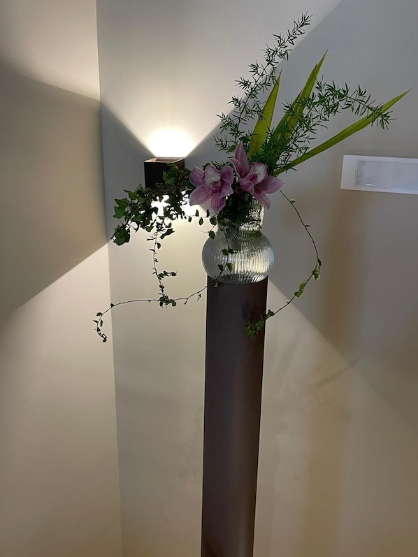 A tall, narrow vase with a textured surface containing pink flowers and green foliage, placed against a plain wall with a light fixture above it. - Ai Cortili (Sant'Egidio del Monte Albino)