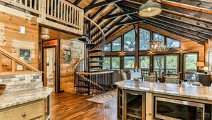 A rustic cabin interior with a wooden staircase, a kitchen area, and large windows. - Lakefront Innisfree | Blue Ridge, GA (Blue Ridge)