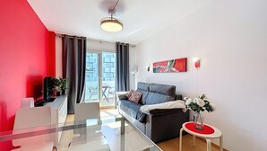 Een moderne woonkamer met een glazen eettafel, een bank, een klein bijzettafeltje met een vaas, en uitzicht op het balkon.