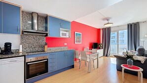Een moderne keuken met blauwe kastjes, een magnetron en een koffiezetapparaat. Er is een eethoek met een tafel en stoelen, en een zithoek met een bank en een raam.