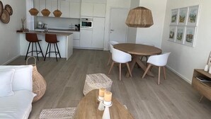 Eine moderne Küche mit weißen Schränken, ein Essbereich mit einem Holztisch und weißen Stühlen, ein Sofa und eine Pendelleuchte.