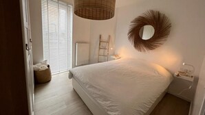 Ein Schlafzimmer mit Bett, Nachttisch, Lampe, Spiegel, Fenster mit Jalousien, einer Leiter und einem Korb.
