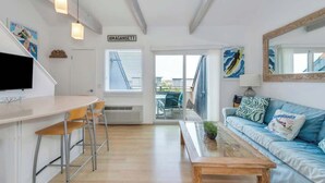 A modern living room with a white sofa, a wooden coffee table, and a TV on a white stand. There is a staircase leading to an upper level, and a door labeled "AMAGANSETT". - Unit 1825: 1-Bedroom with Loft | 2nd Floor-Partial Ocean View | Dunes East (Amagansett)