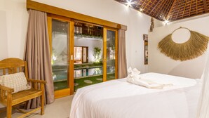 A bedroom with a large bed, a wooden chair, a view through a glass door to an outdoor area, and a thatched wall hanging. - Villa Namu by Alfred in Bali (Seminyak)