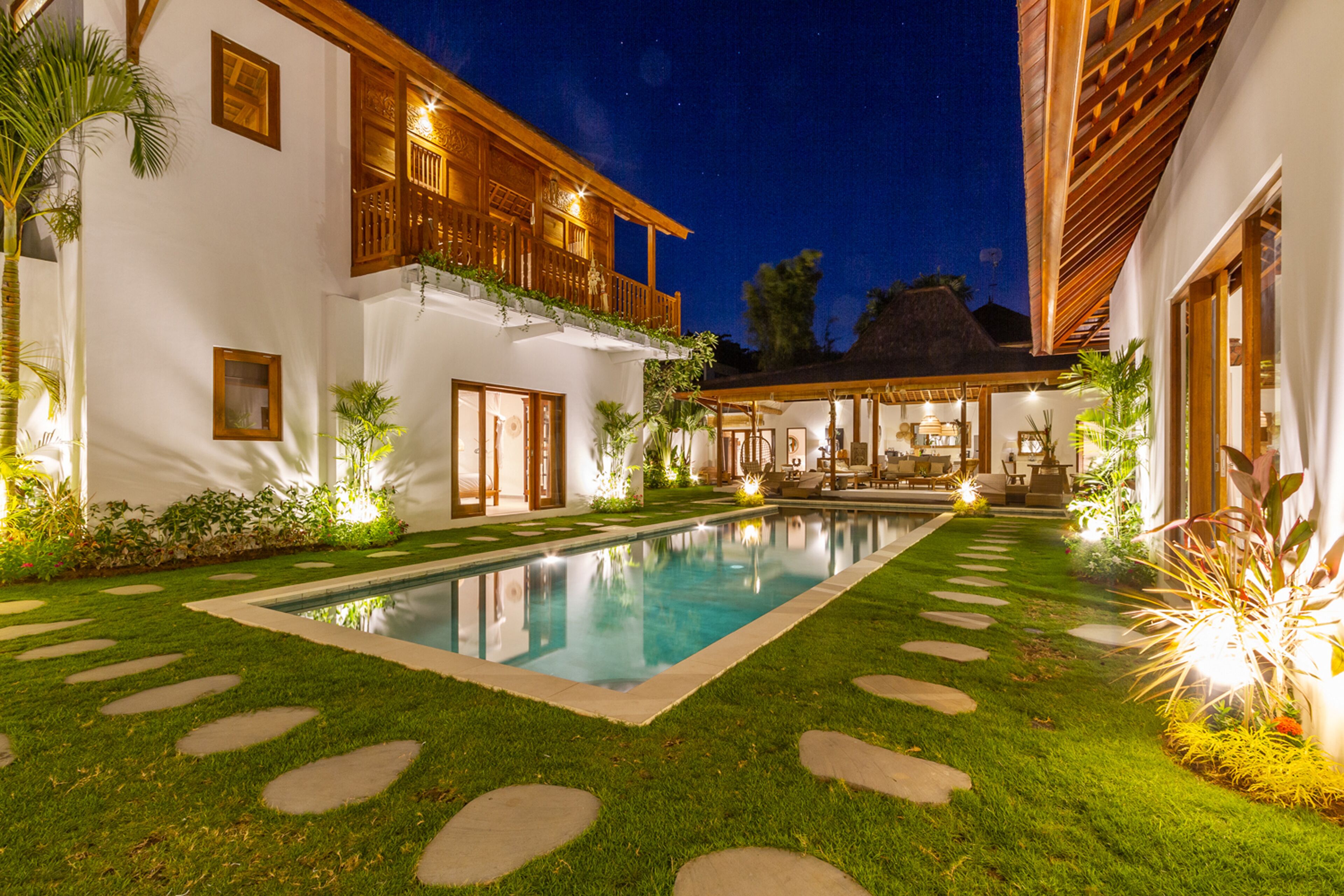 A well-lit pool area with a clear pool, surrounded by a neatly trimmed lawn and stone stepping stones.
