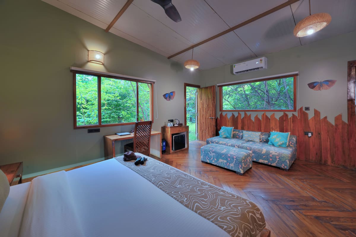 A room with a bed, a desk, a sofa, and large windows with a view of trees.