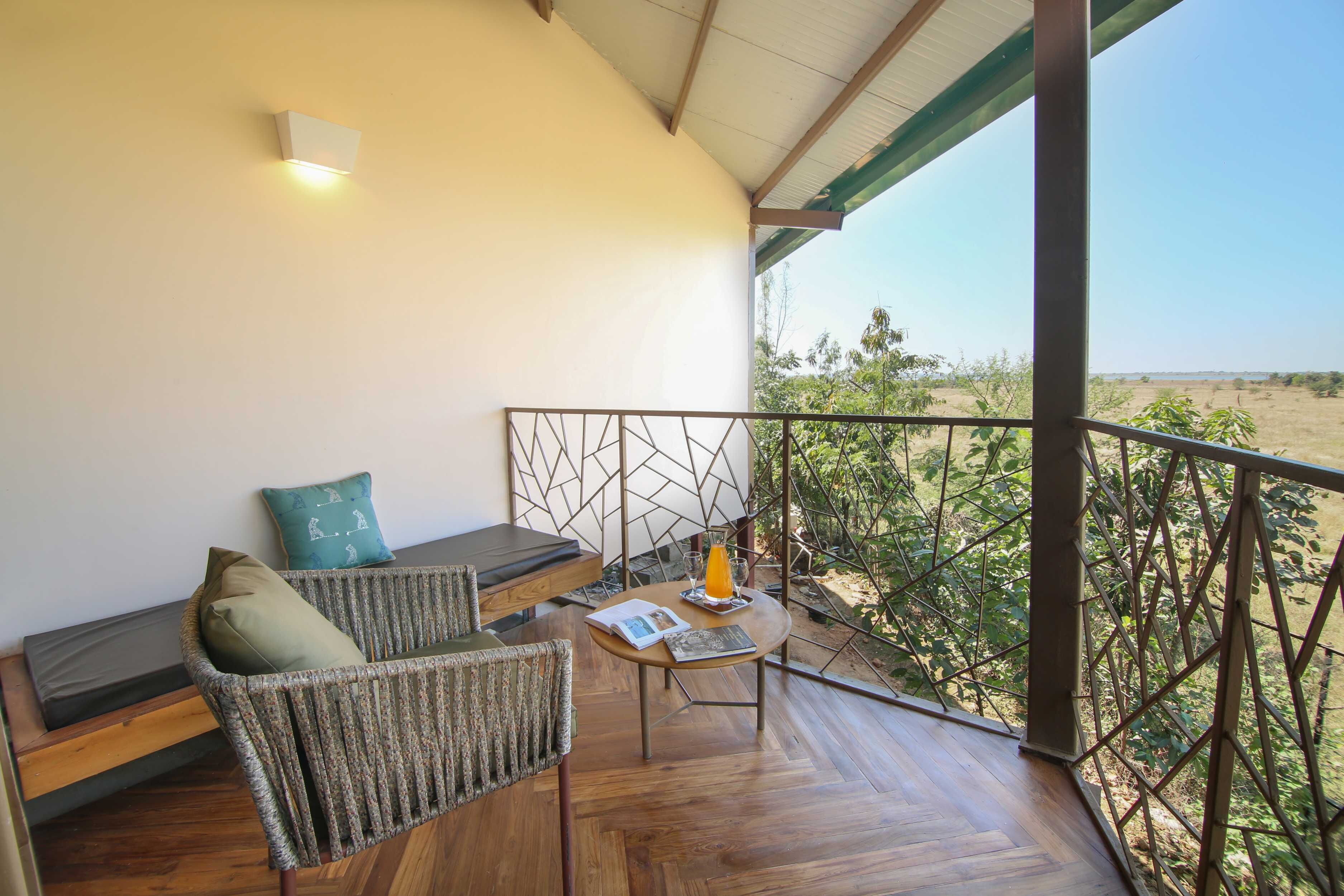 A balcony with a wooden floor, a cushioned seating area with a cushion, a small table with a glass of orange juice, and a view of a natural landscape.