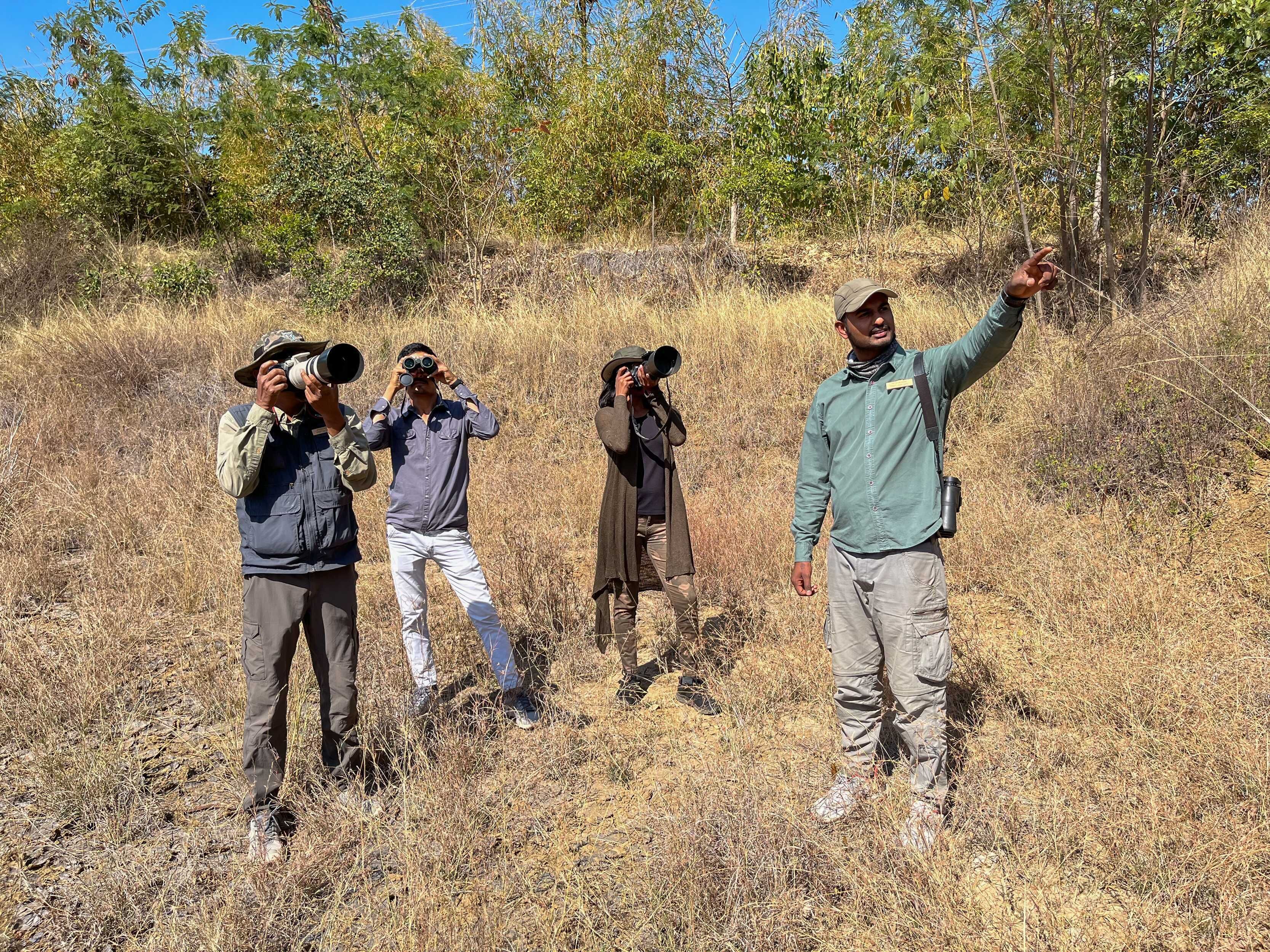 Four individuals in a natural setting with dry grass and trees in the background.