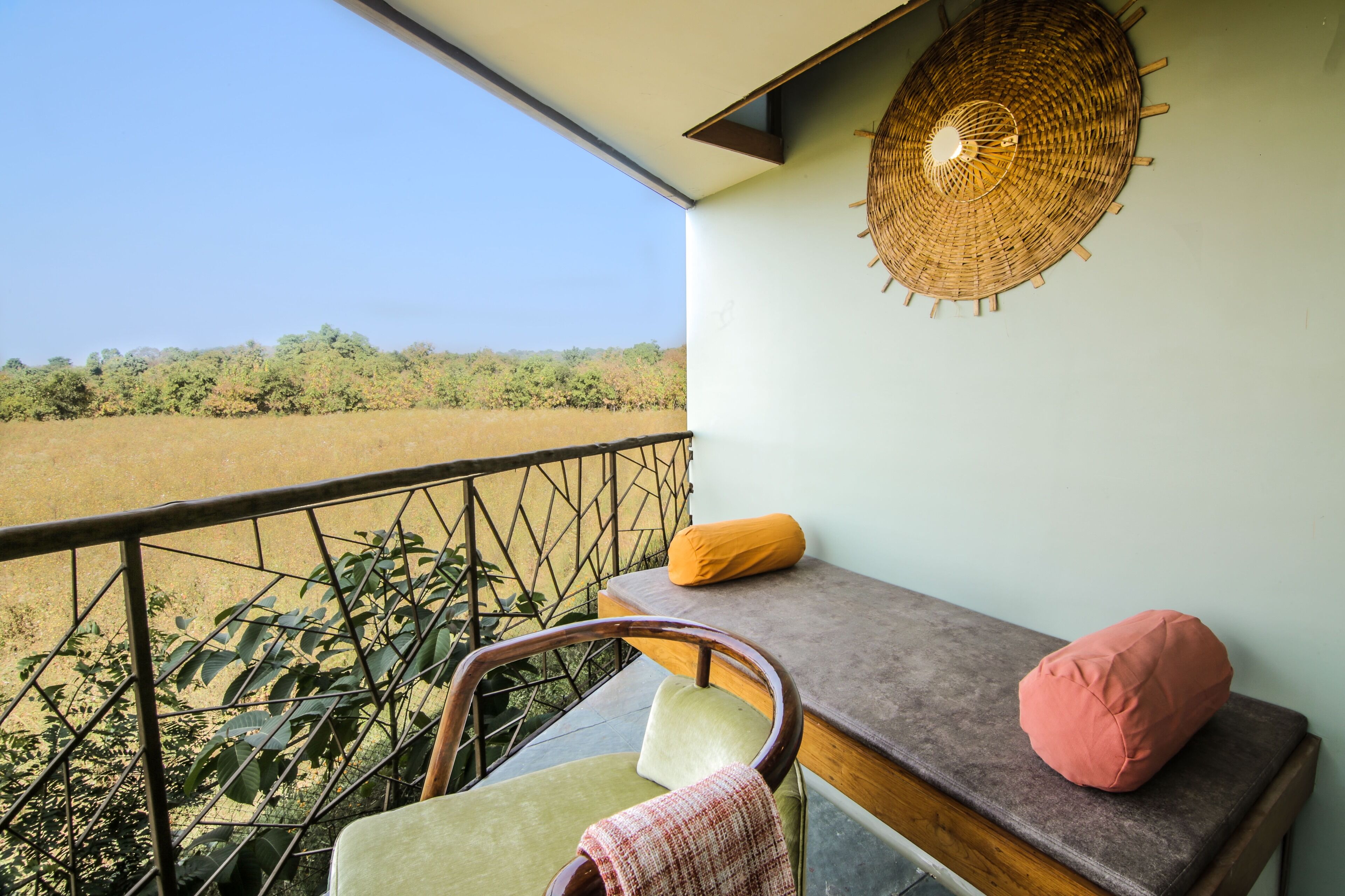 A balcony with a view of a field, a wicker wall hanging, a cushioned bench, and a chair.