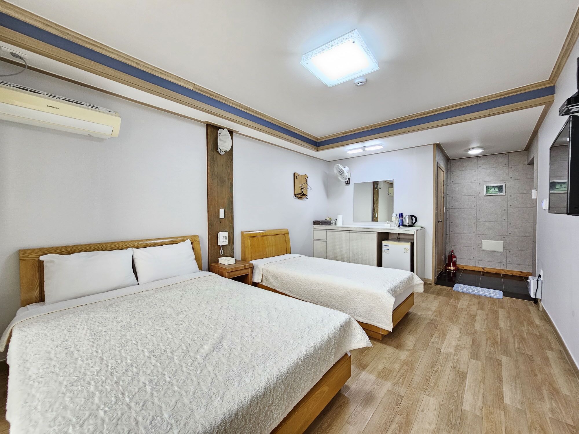 A hotel room with two beds, a desk, a mirror, a small kitchenette, and a ceiling fan.