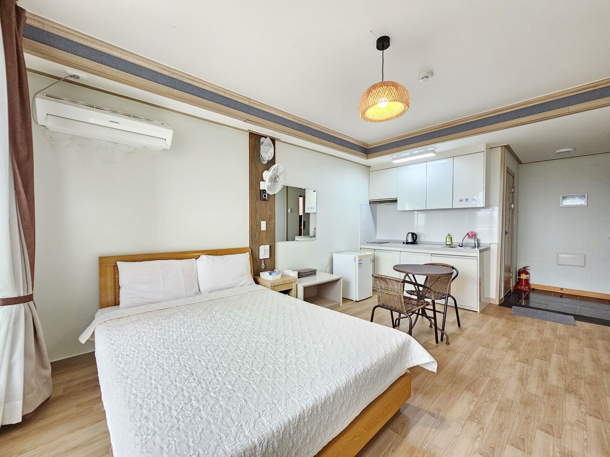 A hotel room with a bed, a small desk, a chair, a kitchenette, and a ceiling fan.