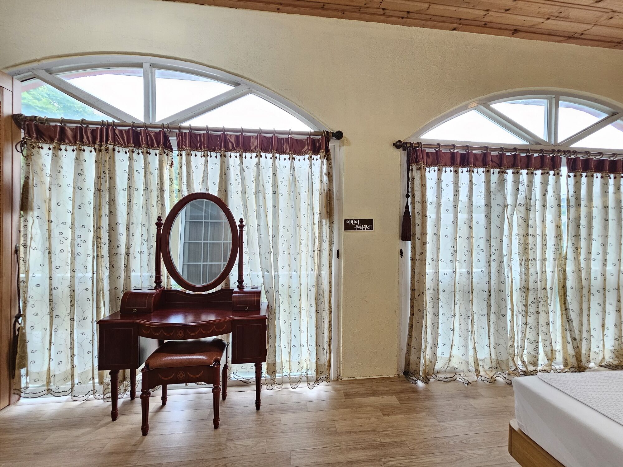 A room with a wooden desk, a mirror, and a bench. There are two windows with curtains.