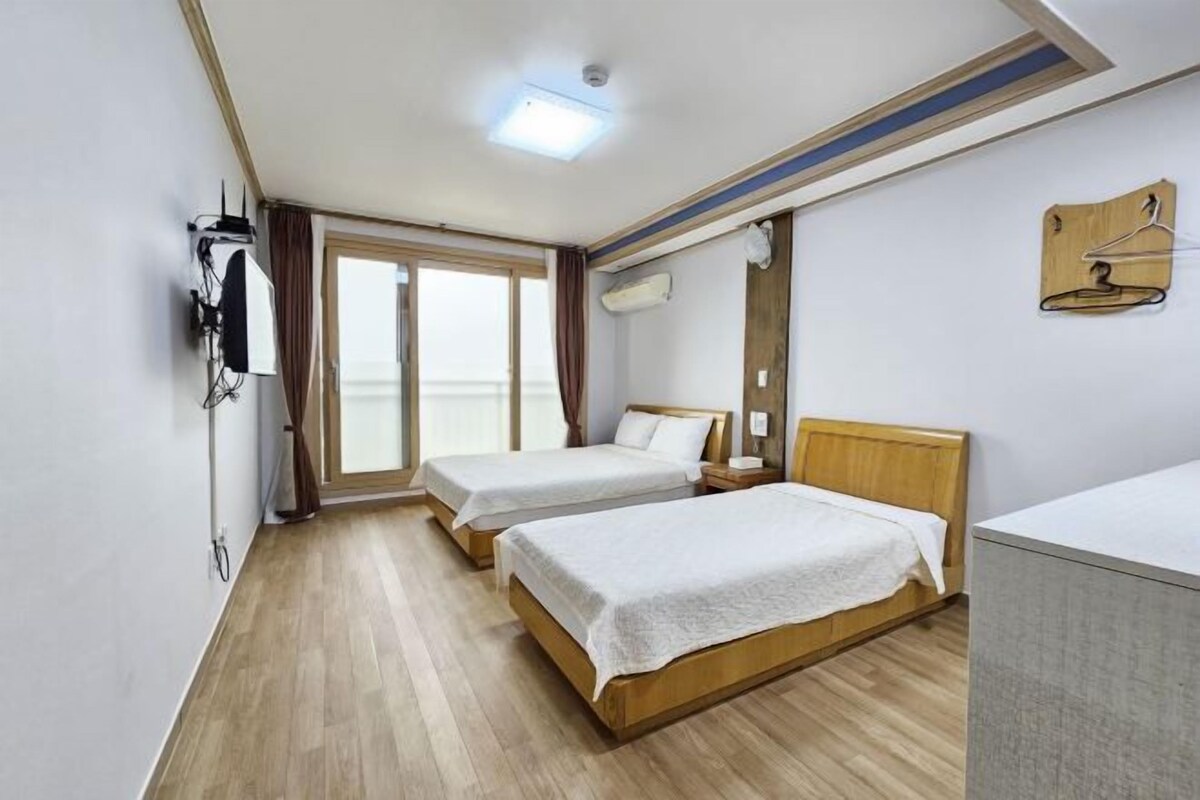 Une chambre d’hôtel avec deux lits simples, un plancher en bois, une grande fenêtre, un bureau et une armoire.