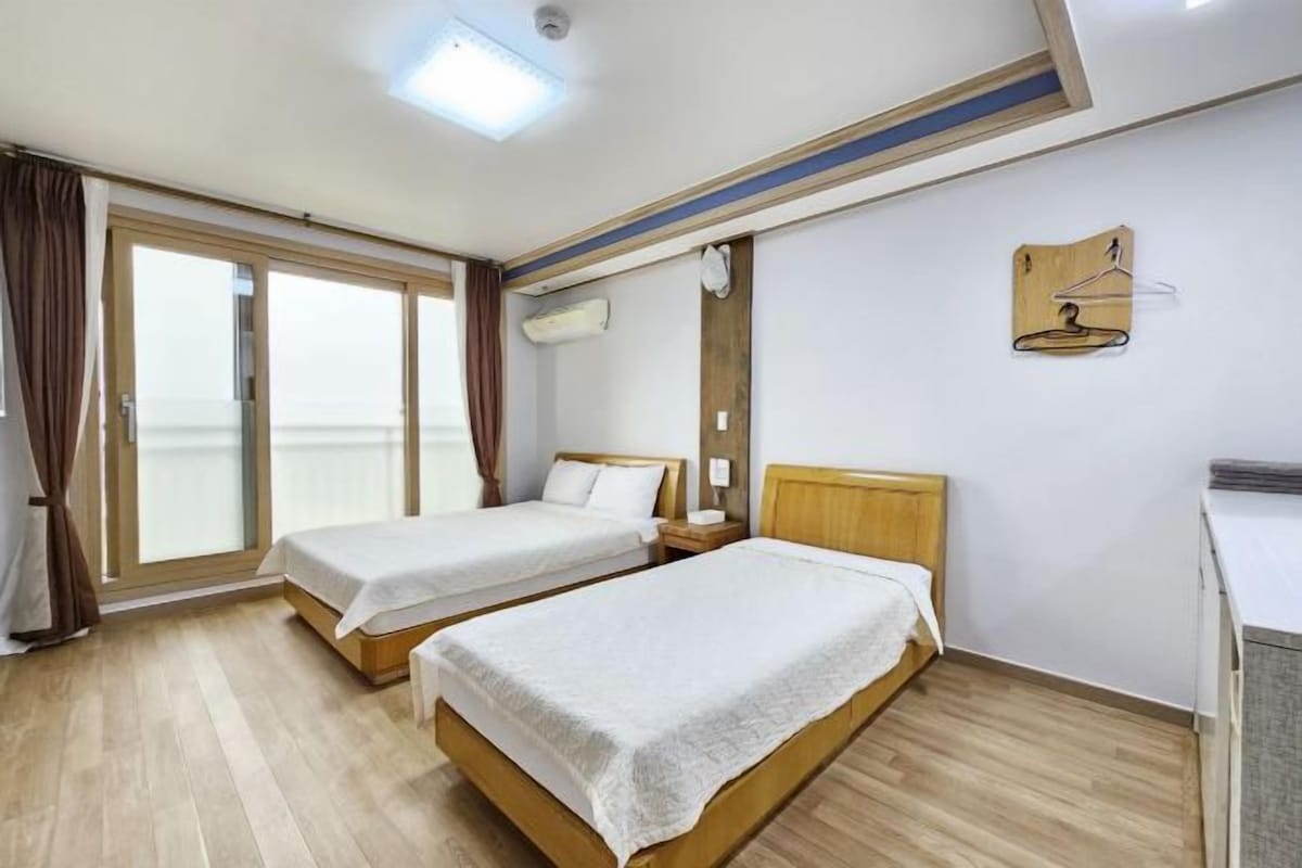 A hotel room with two beds, a wooden floor, a large window with curtains, and a wall-mounted air conditioning unit.
