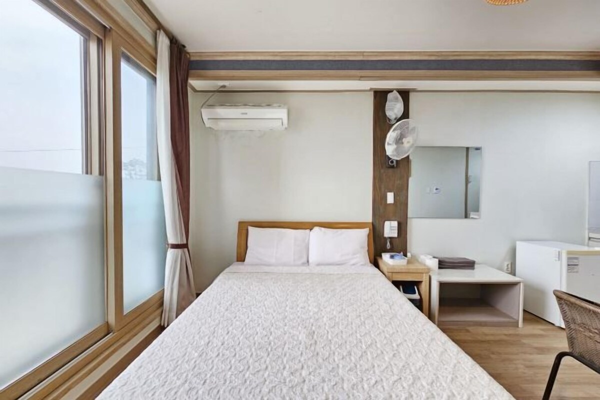 A hotel room with a bed, a desk, a chair, a small table, an air conditioning unit, a mirror, and a window with curtains.