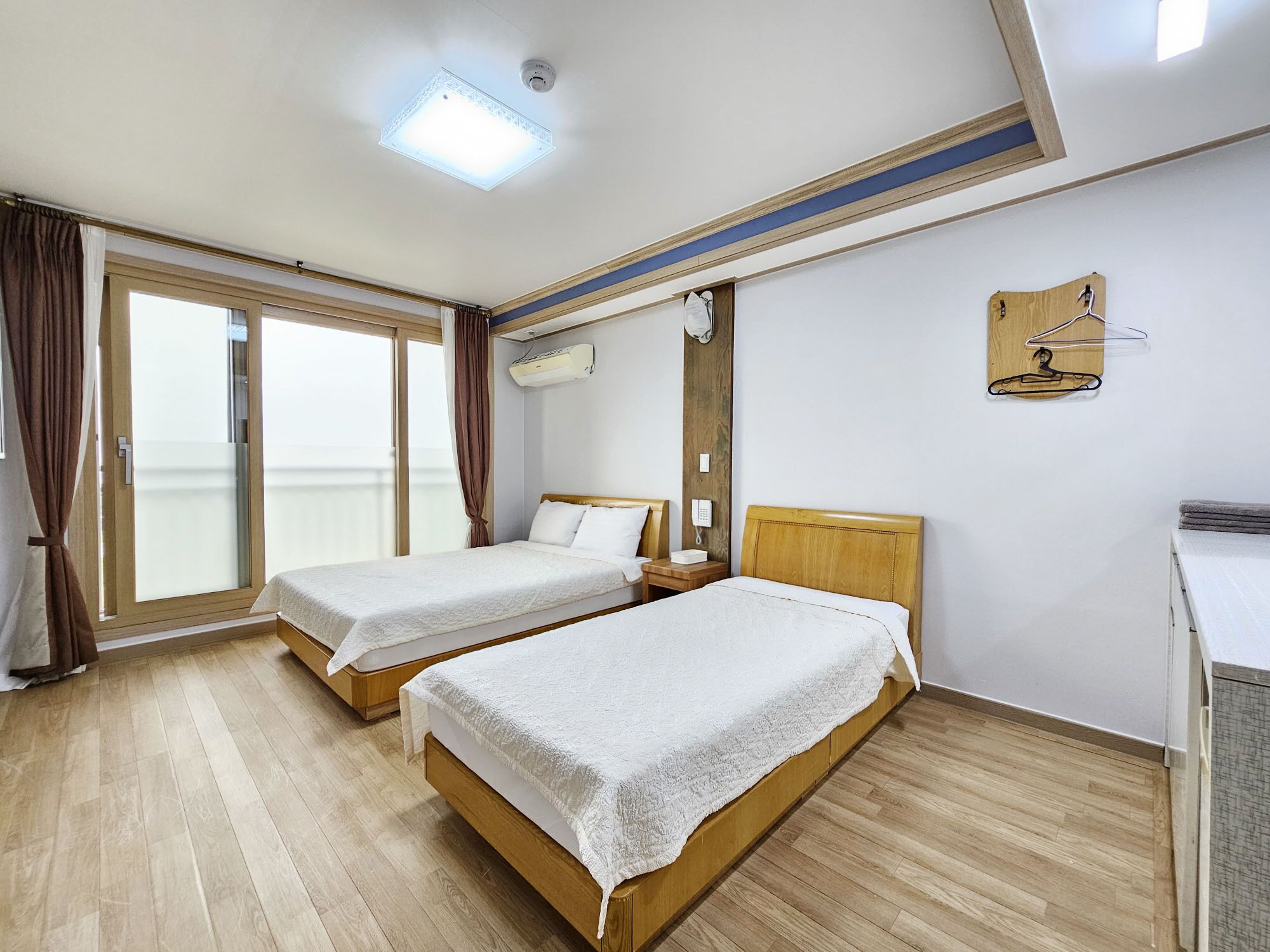 Ein Hotelzimmer mit zwei Betten, einem großen Fenster mit Vorhängen, einer Deckenleuchte, einer Klimaanlage und einem Holzkopfende.