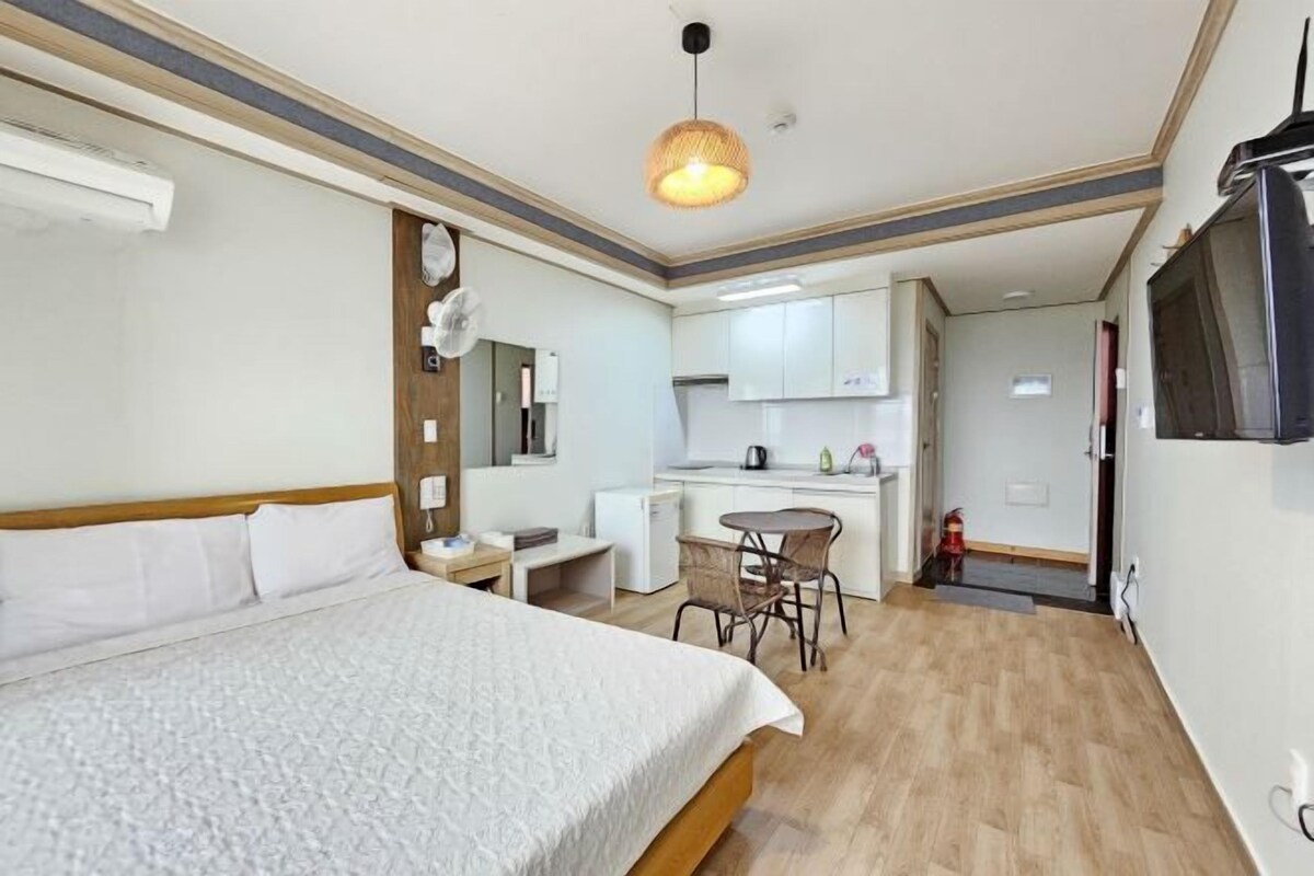 A hotel room with a bed, a small desk, a chair, a kitchenette, and a TV.