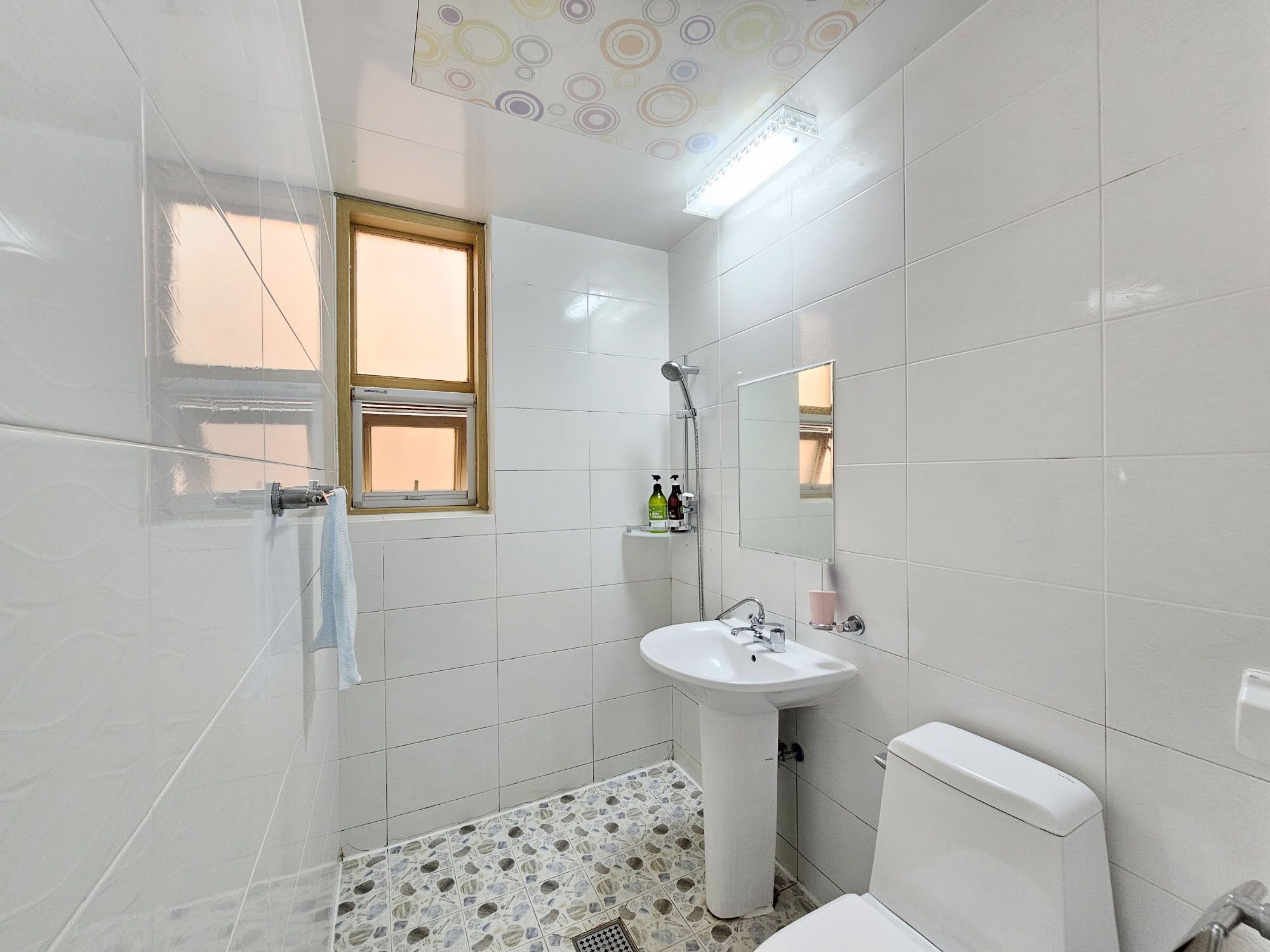 Une salle de bain avec des carreaux blancs, une fenêtre, un lavabo, des toilettes et une douche.