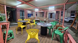 An outdoor patio area with tables and chairs, a covered structure, and a white car parked nearby. - Red Castle Pension (Jeju City)