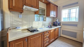 A kitchen with wooden cabinets, a built-in microwave, and a window with blinds. - Red Castle Pension (Jeju City)