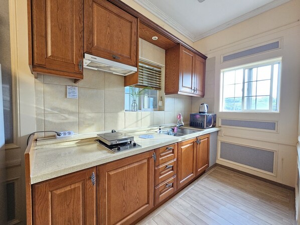 A kitchen with wooden cabinets, a built-in microwave, and a window with blinds. - Red Castle Pension (Jeju City)