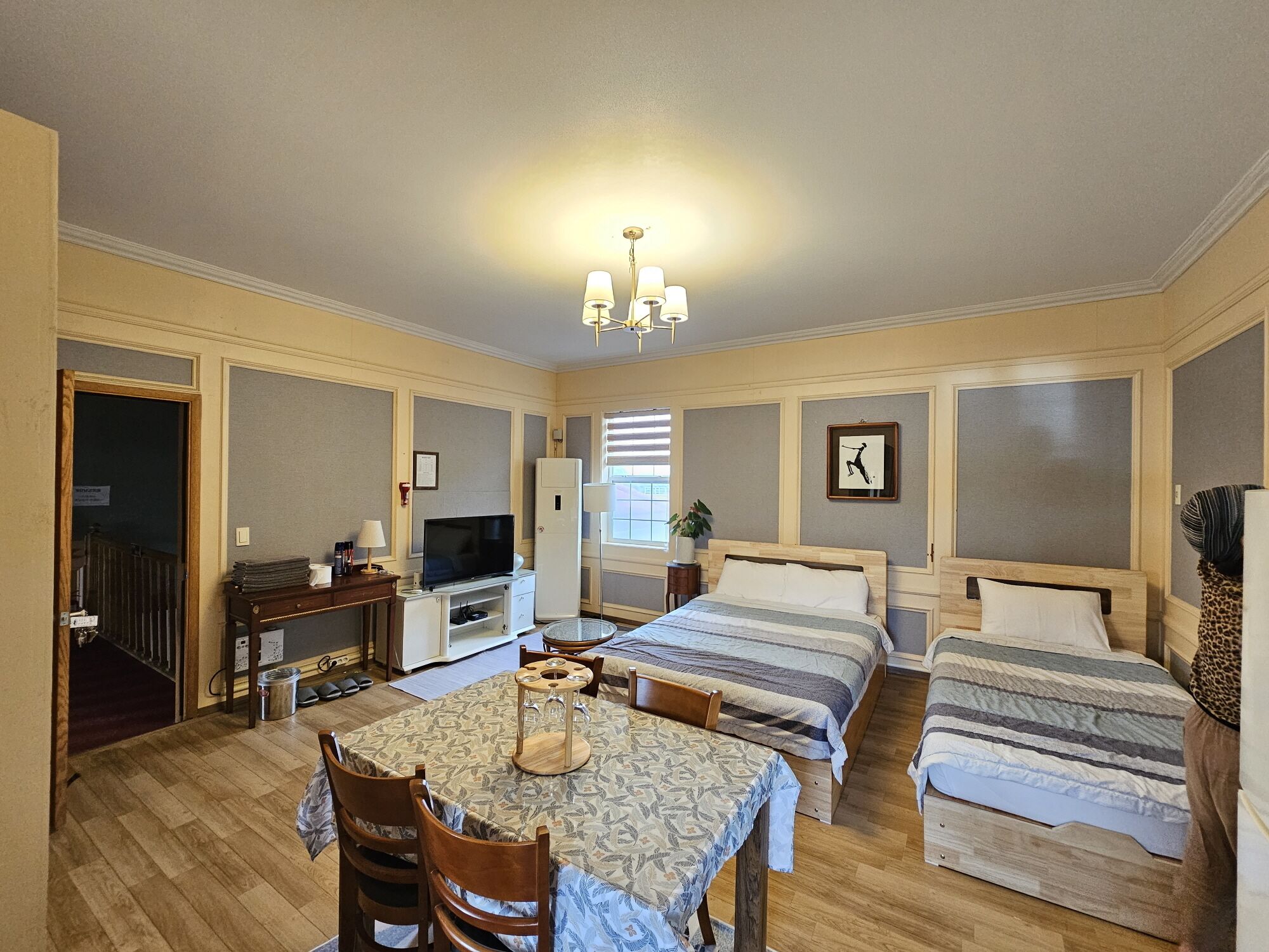 A hotel room with a wooden floor, a dining table with chairs, a TV, a bed, and a chandelier.