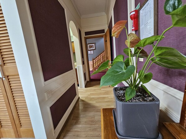 A hallway with a potted plant, wooden flooring, and a staircase in the background. - Red Castle Pension (Jeju City)