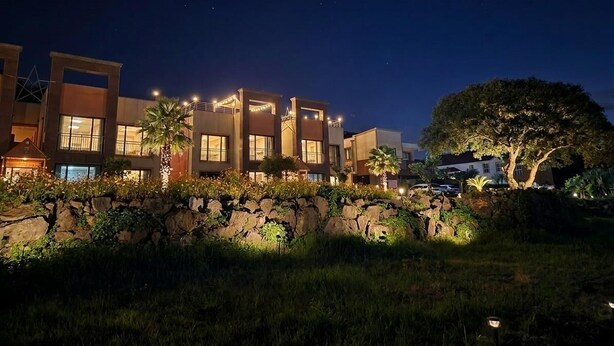 A multi-story hotel with illuminated windows and a stone wall with greenery in front. - Red Castle Pension (Jeju City)