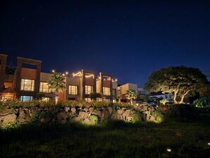 A multi-story hotel with illuminated windows and a stone wall with greenery in front. - Red Castle Pension (Jeju City)