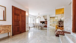 A spacious room with a polished marble floor, a wooden door, a dining table with chairs, and a staircase with a decorative railing. - Villa 'Casa Antonietta' with Private Terrace and Shared Garden (Sant'Anna Arresi)
