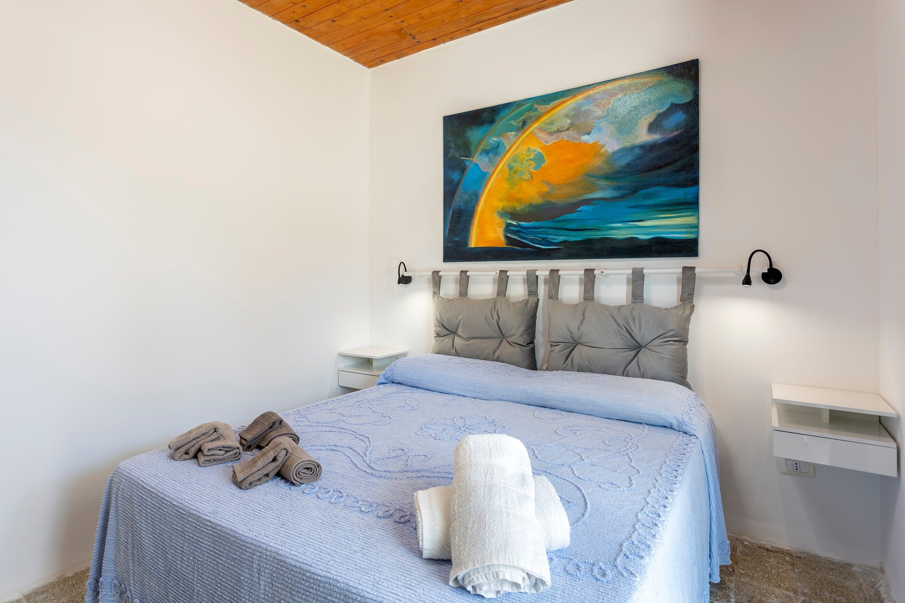 A bedroom with a bed, a painting, a nightstand, a bedside table, and a wall-mounted shelf.