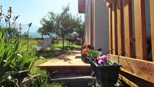 Une terrasse en bois menant à un bâtiment doté d’un toit en pente, entouré de verdure et de fleurs.