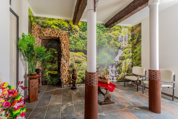 A room with a stone archway, a waterfall mural, a wooden statue, and a seating area with chairs.