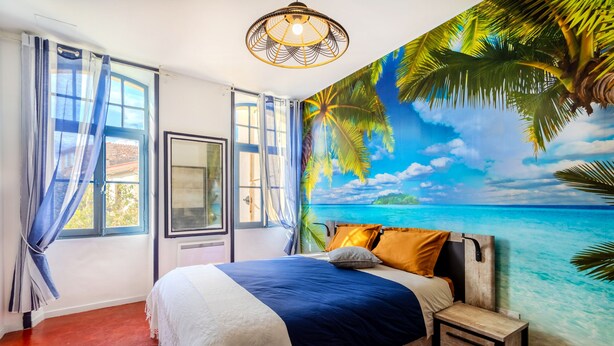 Un dormitorio con cama, mesita de noche, ventana con cortinas y un mural con escena de playa.