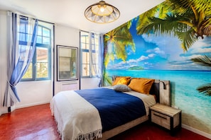 A bedroom with a bed, a nightstand, a window with curtains, and a mural of a beach scene.